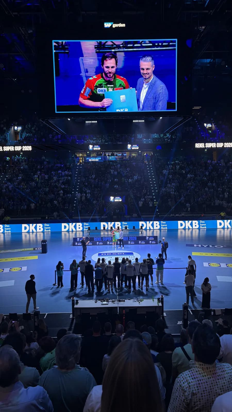 Videowürfel im SAP Garden zeigt die Übergabe einer überdimensionalen DKB-Karte an einen Handballspieler